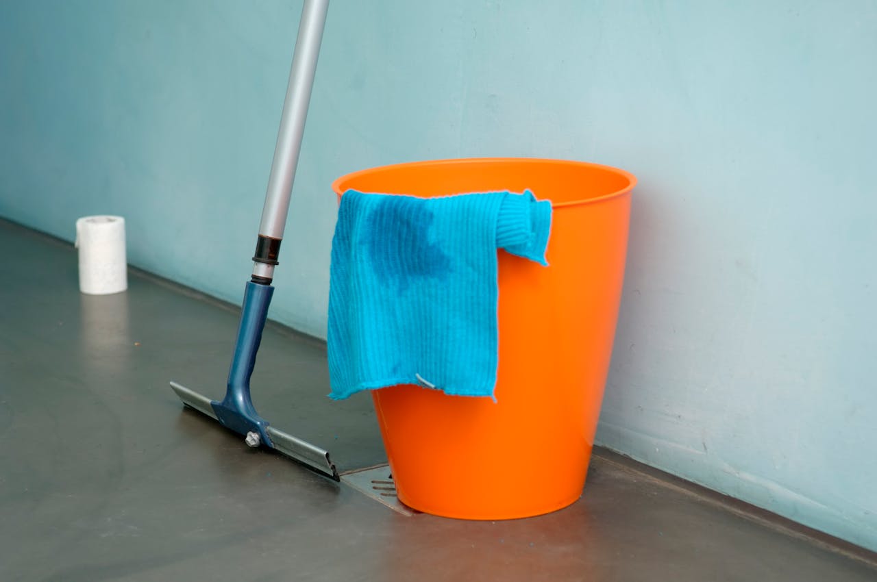 Close-up of cleaning supplies including orange bucket, mop, and blue rag indoors.